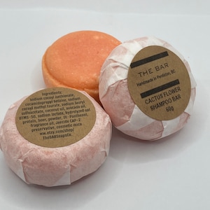 May include: Three pink shampoo bars wrapped in white paper with brown labels. The label on the front bar reads "THE BAR Handmade in Penticton, BC CACTUS FLOWER SHAMPOO BAR 60g". The label on the back bar lists the ingredients.