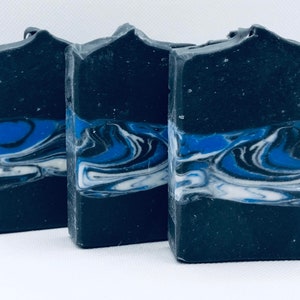 May include: Three rectangular bars of handmade soap. The soap is mainly black with a blue and white marbled design across the centre. The top edges of the soap have a slightly uneven, textured appearance.