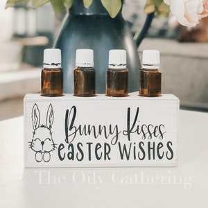 May include: White wooden essential oil holder with a bunny illustration and the text "Bunny Kisses Easter Wishes". The holder has four slots for essential oil bottles. The holder is sitting on a white surface in front of a bouquet of pink flowers.
