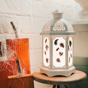 May include: White ceramic lantern with a cut-out design of leaves. The lantern is lit and sitting on a wooden stand with black metal legs.