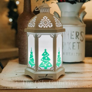 May include: White lantern with a cut-out design and green Christmas tree images. The lantern is lit and has a "Young Living" logo on the bottom.