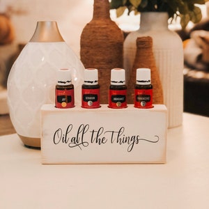 May include: White wooden essential oil holder with the text "Oil all the things" in black script. Four small glass bottles with red labels are in the holder. The labels read "Joy", "Geranium", "Abundance", and "Frankincense".