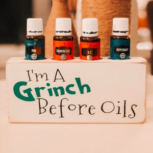May include: A white wooden box with the text "I'm a Grinch Before Oils" in green and black lettering. The box holds four small glass bottles with white lids, labelled "Pine", "Frankincense", "R.C." and "Peppermint".