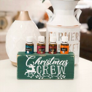 May include: A green wooden box with the words "Christmas Crew" and a white reindeer design. Four small glass bottles with labels for "Peppermint", "Purification", "Thieves", and "Orange" are displayed in the box.