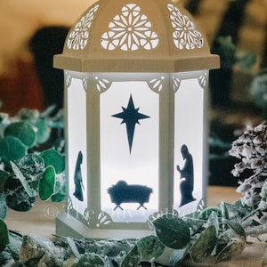 Może przedstawiać: Biała metalowa latarnia z wyciętym wzorem przedstawiającym gwiazdę, żłóbek i dwie postacie. Latarnia jest oświetlona od wewnątrz, dając ciepłe światło.