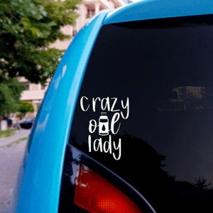 May include: A blue car with a white vinyl sticker on the back window that says "crazy oil lady" with a small image of a bottle of oil.