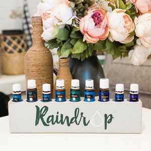 Może przedstawiać: Drewniane pudełko z napisem "Raindrop" i namalowanym symbolem kropli deszczu. Pudełko zawiera dziesięć małych szklanych butelek z białymi zakrętkami, wypełnionych niebieskim i fioletowym płynem. Butelki są ułożone w rzędzie.