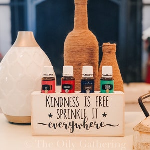 May include: White wooden essential oil holder with the text "Kindness is free sprinkle it everywhere" and a star on either side of the word "sprinkle". The holder has five slots for essential oil bottles. Four bottles are in the slots, two red, one blue, and one green. The holder is on a white surface.