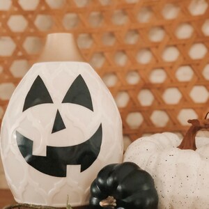 May include: White ceramic pumpkin-shaped diffuser with a black jack-o'-lantern face. The diffuser is sitting on a wooden surface with a small black and white pumpkin and a small black pumpkin beside it.