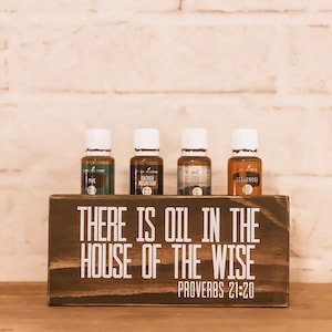 May include: A wooden sign with the text "There is oil in the house of the wise, Proverbs 21:20". Four small brown glass bottles with white lids are sitting on top of the sign. The bottles are labelled "Pine", "Sacred Mountain", "Sacred Angel", and "Cedarwood".