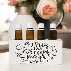 May include: White wooden box with four small brown glass bottles with white lids. The box has the text "This too shall pass" in black cursive lettering with decorative swirls. The text "The Oily Gathering" is in black lettering at the bottom of the box.