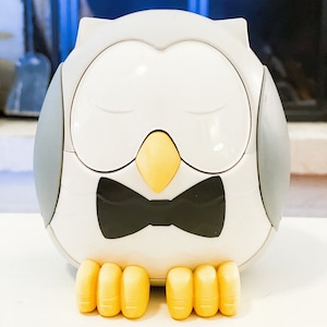 May include: A white owl-shaped ceramic piggy bank with a black bow tie and yellow feet. The owl has its eyes closed.