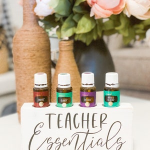 May include: A white wooden block with the words "Teacher Essentials" in black script holds four essential oil bottles. The bottles are labeled "Thieves," "Clarity," "En-R-Gee," and "Peace & Calming." Decorative bottles wrapped in twine and a bouquet of flowers are in the background.