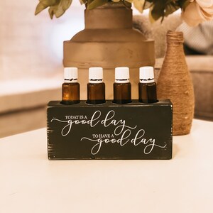 Puede incluir: Un soporte de madera negro para aceites esenciales con el texto "Today is a good day to have a good day" en letras blancas. El soporte tiene cuatro ranuras para botellas de aceites esenciales, y hay cuatro botellas de vidrio marrón con tapas blancas en las ranuras.