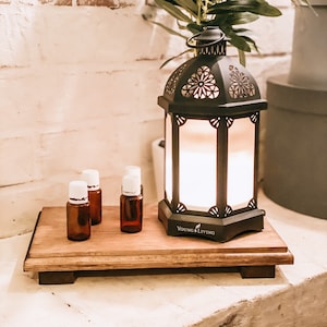 May include: A wooden tray with four small glass bottles and a black metal lantern with a white light inside. The lantern has a decorative design and says "Young Living" on the bottom. The tray is sitting on a white brick surface.