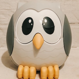 Może przedstawiać: Szary pojemnik w kształcie sowy z dużymi czarnymi oczami i żółtym dziobem. Pojemnik ma fakturowaną powierzchnię i stoi na białym tle.