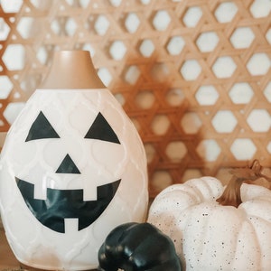 May include: White ceramic pumpkin-shaped diffuser with a black jack-o'-lantern face. The diffuser is surrounded by three small pumpkins, one black, one white with black speckles, and one white with brown stem.