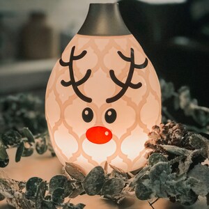May include: A white ceramic essential oil diffuser with a reindeer design. The diffuser has a silver top and is lit up, casting a warm glow. The diffuser is surrounded by a garland of green leaves with a dusting of white snow.