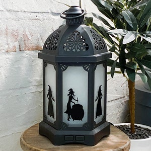 Może przedstawiać: Czarna metalowa latarnia z białym, matowym panelem szklanym przedstawiającym sylwetkę trzech czarownic. Latarnia ma ozdobny, skomplikowany wzór i uchwyt na górze.