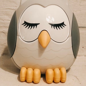 May include: A white owl-shaped sleep sound machine with gray wings and yellow feet. The owl has closed eyes with eyelashes and a yellow beak.