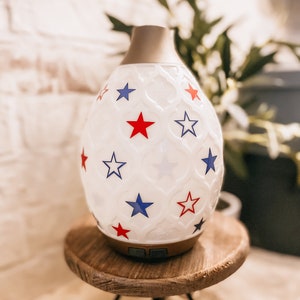 Puede incluir: Difusor de aceites esenciales de cerámica blanca con un patrón de estrellas en rojo, azul y blanco. El difusor está sentado en un soporte de madera.