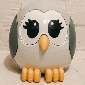 Może przedstawiać: Biała zabawka w kształcie sowy z szarymi skrzydłami, czarnymi oczami z rzęsami i żółtym dziobem. Sowa ma cztery żółte łapy.