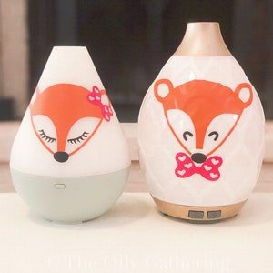 May include: Two ceramic essential oil diffusers with a fox design. The smaller diffuser is light green with a white fox face and pink bow. The larger diffuser is white with a gold top and a fox face with a pink bow tie.