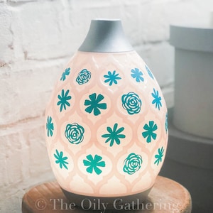 Op de afbeelding: Een witte keramische etherische olieverdamper met een zilveren bovenkant en een turquoise bloemenpatroon. De diffuser staat op een houten tafel.