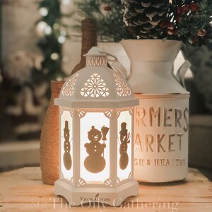 Op de afbeelding: Witte keramische lantaarn met een uitgesneden ontwerp met drie sneeuwpop-silhouetten. De lantaarn is verlicht en gloeit warm. De lantaarn heeft de tekst "Young Living" op de onderkant.
