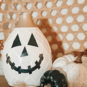 May include: White ceramic diffuser shaped like a jack-o'-lantern with a black smiling face. The diffuser is surrounded by three small white pumpkins with black speckles and a black pumpkin.