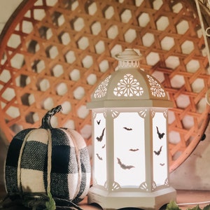 Op de afbeelding: Witte lantaarn met een uitgesneden ontwerp van vleermuizen. De lantaarn is verlicht en geeft een warm gloed. Een zwart-wit geruite pompoen staat voor de lantaarn.