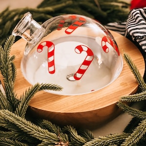 May include: A clear glass diffuser with a wooden base, decorated with red and white candy cane designs. The diffuser is surrounded by green pine branches.