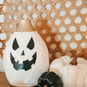 Puede incluir: Difusor de cerámica blanco con forma de calabaza de Halloween con una cara negra. El difusor está sobre una superficie de madera con una calabaza blanca y una calabaza negra.