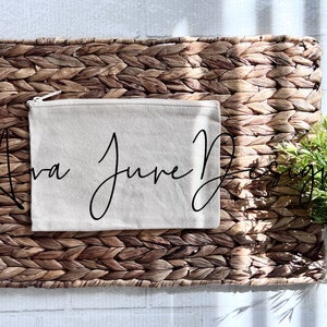 May include: A beige canvas zippered pouch with the text "Ava June Designs" written in black cursive script. The pouch is sitting on a woven brown wicker mat.