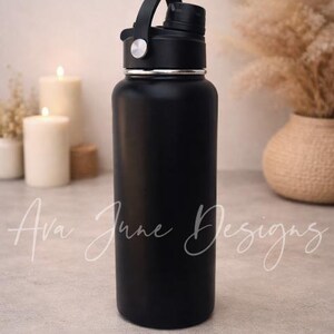 May include: A matte black water bottle with a black handle and a silver rim. The bottle is cylindrical and has a black cap. The background includes candles, pampas grass, and the text "Ava June Designs".