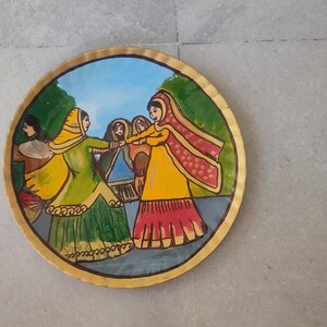 K&ouml;nnte beinhalten: Ein runder, bemalter Teller mit goldfarbenem Rand zeigt eine Szene mit vier Frauen in bunten Kleidern, die in einem Kreis tanzen. Die Frauen tragen traditionelle indische Kleidung, darunter Saris und Dupattas. Der Hintergrund ist eine gr&uuml;ne und blaue Landschaft.