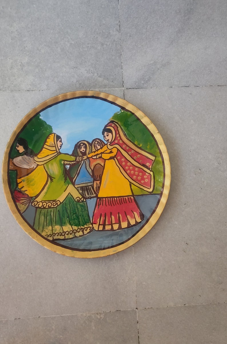 K&ouml;nnte beinhalten: Ein runder, bemalter Teller mit goldfarbenem Rand zeigt eine Szene mit vier Frauen in bunten Kleidern, die in einem Kreis tanzen. Die Frauen tragen traditionelle indische Kleidung, darunter Saris und Dupattas. Der Hintergrund ist eine gr&uuml;ne und blaue Landschaft.