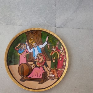 K&ouml;nnte beinhalten: Ein runder Holzteller mit einer bemalten Szene von Menschen in traditioneller Kleidung. Die Szene zeigt eine Frau, die Wolle an einem Spinnrad spinnt, einen Mann mit einem gelben Turban und zwei Frauen in bunten Kleidern. Der Teller hat einen goldenen Rand.