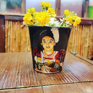 May include: A black metal planter with a painted image of a woman wearing a traditional Indian outfit and jewelry. The planter is filled with yellow flowers.