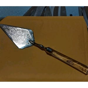 May include: A silver trowel with an ornate, engraved blade and a twisted metal handle. The handle transitions into a clear, rectangular grip filled with a light-colored material. The trowel is designed for decorative or ceremonial use.