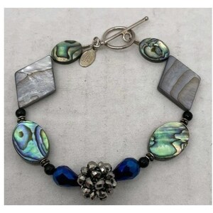 May include: A silver-toned bracelet featuring iridescent abalone shell ovals and diamond-shaped pieces. The bracelet also includes dark blue beads and a cluster of silver-toned beads. The clasp is a toggle style.