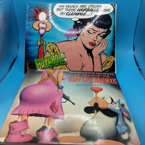 May include: A stack of comic books with colorful illustrations. The top book features a crying woman with a thought bubble that reads, "HIS KISSES ARE DREAMY... BUT THOSE HAIRBALLS DOWN MY CLEAVAGE...!" The bottom book is titled "THE NIGHT OF THE MARY KAY COMMANDOS."