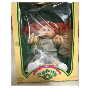 Puede incluir: Muñeca Cabbage Patch Kids vintage de 1985, en su caja original. La muñeca tiene pelo rojo de hilo y ojos azules, vestida con un atuendo gris y amarillo. La caja muestra el logotipo de Cabbage Patch Kids y el lema "I'm unique, I'm adoptable!"