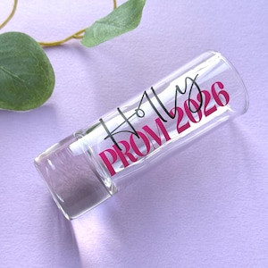 May include: A clear glass shot glass with the text "Holly PROM 2026" in black and pink. The shot glass is on a light purple surface with green leaves in the background.