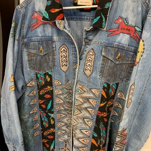 May include: A light blue denim jacket with colourful painted designs. The jacket features red horses, feathers, and geometric patterns. The collar and front panels have black fabric with feather patterns. The brand name "Bear Classics" is visible.