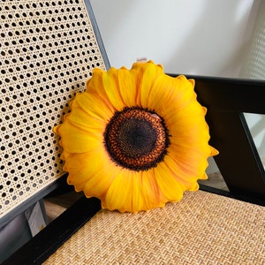 以下が含まれることがあります： 茶色の芯を持つ黄色のひまわりのクッション。クッションは籐製の椅子の上に置かれています