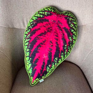 May include: A red, green and black leaf-shaped cushion. The cushion is made of fabric and has a realistic leaf design.