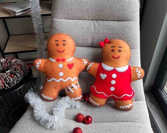 May include: Two gingerbread person-shaped cushions, one in a red dress with white polka dots and a white collar, and the other in a brown gingerbread man costume with a red bow tie. The cushions are sitting on a light grey sofa.