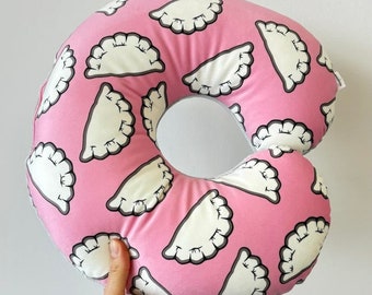 May include: A pink donut-shaped pillow with a white and gray pattern of pierogies. The pillow is soft and plush, perfect for cuddling or supporting your neck.