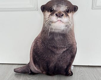 May include: A plush toy shaped like an otter. The otter is brown and gray with a white chest and a black nose. It has a realistic appearance and is sitting on a white surface.